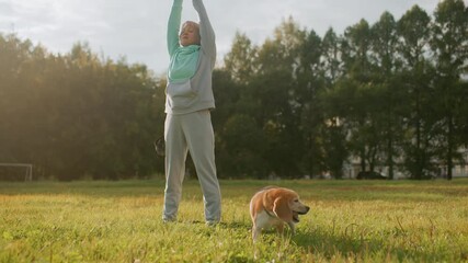 Tamer in mint and grey tracksuit standing on grassy park raising hands upward eyes closed performing morning stretching exercise beside beagle dog under soft sunlight
