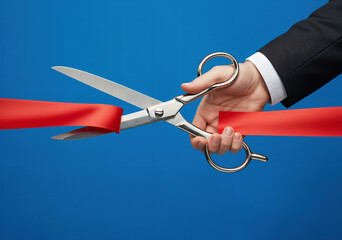 A hand in a suit cutting a wide red ribbon with large silver scissors against a vibrant blue backdrop. Represents grand opening, celebration, and new beginnings