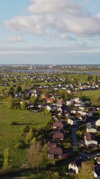 Vertical drone pull out over Katlakalns residential area shows houses, fields, tree lines, Riga skyline, scattered clouds and soft light, urban expansion scene, slow motion, springtime