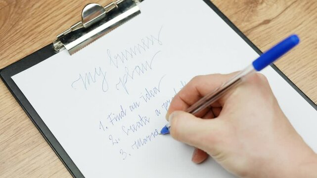 Close-up of a hand writing a business plan on a clipboard using a blue pen with phrases like "Find an idea" and "Search market" are visible on the list, suggesting a creative planning session.