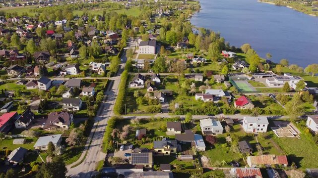 Katlakalns residential area with colorful houses, garden plots, roads, and Daugava river shoreline, tree-lined streets, and distant urban skyline, aerial view, slow motion, springtime