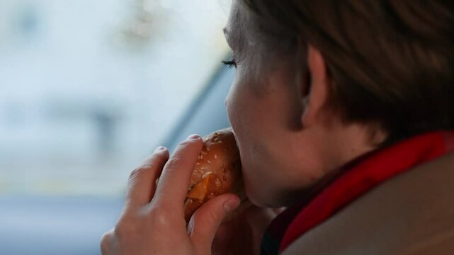 Famished lady takes bite of burger to quell hunger sitting in vehicle on cloudy day. Burger delivered for lady eating burger in vehicle. Lady indulging in unhealthy burger while driving in vehicle