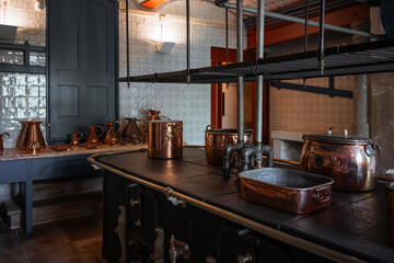 Low-key interior of a historic kitchen featuring a vintage cast iron stove with aged copper pots and pans, surrounded by tiled walls and traditional dark cabinetry under moody lighting