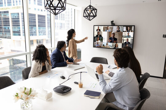 Group of multi-ethnic shareholders gathered around conference table in modern office, participating in videoconference call with diverse partners displaying in screen. Videocall app usage, business