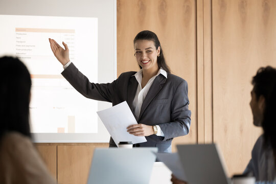Young 30s European business woman, female trainer or corporate coach presenting visual data, pointing on projected slide displaying analytical and financial information to diverse seminar participants