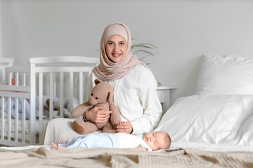 Happy Muslim mother in hijab with teddy bear and cute little baby sleeping on bed at home