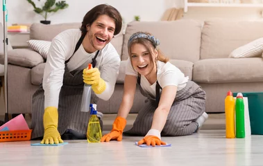 Gordijnen Young happy couple in rubber gloves cleaning floor in their apartment and smiling at camera, washing flat with spray detergent and rag © Prostock-studio
