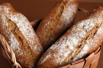 Wicker basket with fresh rye baguettes on brown background, closeup