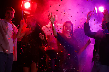 Group of young people with falling confetti dancing at party in night club