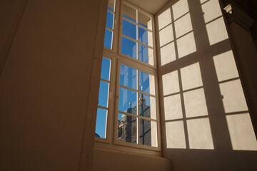 Fototapeta premium Sunlight streams through a multi-pane window, casting geometric shadows onto a smooth, beige interior wall. The view includes a ornate rooftop structure and a cross-topped spire of Berlin Cathedral. 
