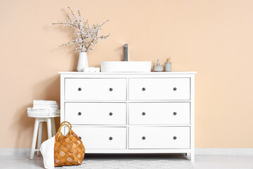 Vase with blooming branches and sink on chest of drawers near beige wall in interior of bathroom