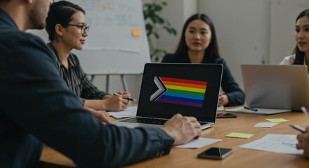 Diverse team in a modern office meeting discussing LGBTQ+ inclusion, with a laptop displaying the Progress Pride flag.