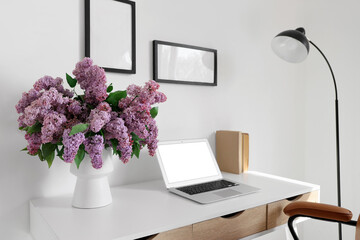 Vase with lilac flowers and blank laptop on desk in office