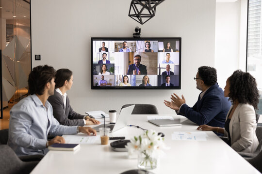 Group of diverse businesspeople displayed on screen during videocall while multiethnic teammates listening speech of boss seated at conference table in modern boardroom. Business virtual meeting event