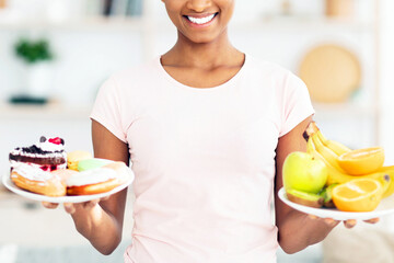 Lovely black lady holding plates with fruits and sweets, looking at unhealthy pastries, tempted to...