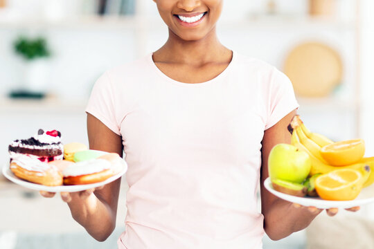 Lovely black lady holding plates with fruits and sweets, looking at unhealthy pastries, tempted to eat them at home. Beautiful woman trying to keep healthy diet, resisting urge to have junk food