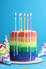 Delicious rainbow birthday cake with burning candles, party hats and confetti on light blue...