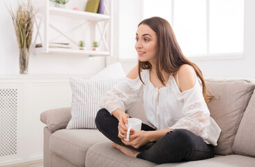 Obraz premium Happy interested girl at home, watching TV, drink coffee on sofa, astonished face