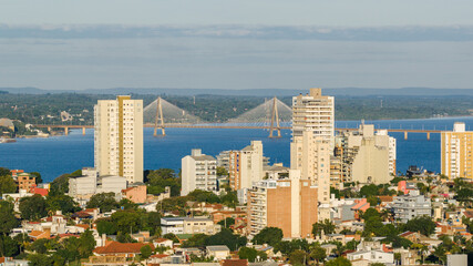 Panoramic view of the city of Encarnación, Paraguay, with modern buildings in the foreground and the San Roque González international bridge crossing the Paraná River in the background.