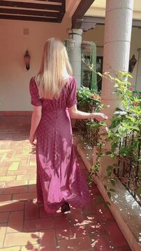 Vertical video shows a young woman with long blonde hair, dressed in a long, red dress, walking in slow motion, facing backwards, on the terrace of a Spanish-style villa.  