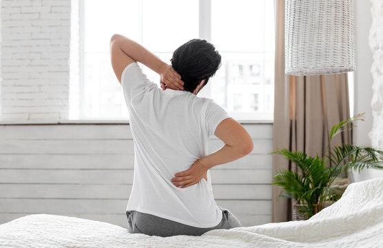 Unrecognizable guy suffering from back pain in bedroom, waking up in morning, sitting on bed and touching his neck and lower back with both hands, wearing pajamas, copy space, back view