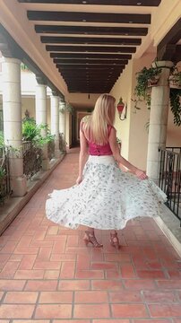 Vertical video shows a young woman with long blonde hair, dressed in a long white skirt, spinning in slow motion on the terrace of a Spanish-style villa.