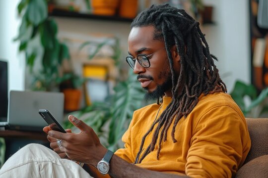 young man with dreadlocks and glasses sitting and using smartphone in cozy room with plants and laptop in background