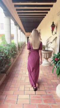 Vertical video shows a young woman with long blonde hair, dressed in a long, red dress, slowly walking, facing backwards, on the terrace of a Spanish-style villa.  