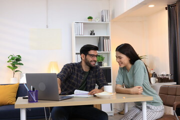 Business couple working, communicating while sitting at the home office desk together. Cheerful couple, calm asian woman and caucasian man working at home with laptop together.