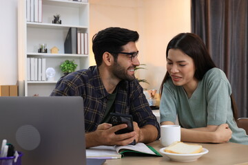 Business couple working, communicating while sitting at the home office desk together. Cheerful couple, calm asian woman and caucasian man working at home with laptop together.