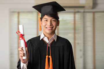 Asian young graduate wearing black cap and gown holds diploma with red ribbon, smiling confidently...