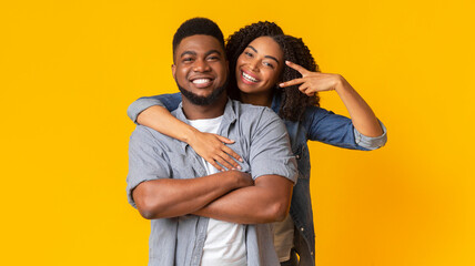 Fototapeta premium Portrait of millennial afro couple embracing and posing at camera, woman showing peace gesture, yellow studio background, panorama