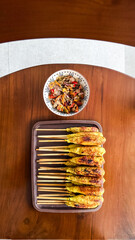 Indonesian satay lilit is served on skewers with traditional sambal matah. It is a delicious Balinese dish presented on a wooden table in a top-down view.