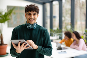 App for knowledge. Cheerful european guy students using tablet for video lesson, studying with friends in classroom, ladies sitting at table on background