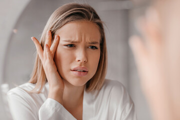 Mirror reflection of upset young lady looking at mirror and touching her forehead, having oily or...