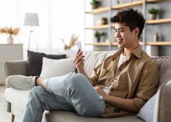 Obraz premium Cool Gadget And Application. Portrait of young smiling asian man holding mobile phone, typing sms message, sitting on the sofa in living room. Guy browsing internet, surfing web, using app, free space