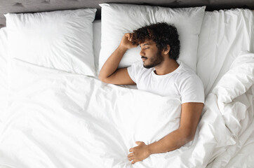 Top view of handsome bearded young hindu guy in white t-shirt resting at home, sleeping alone in...