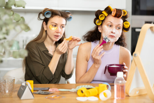 Female friends enjoying moments of beauty routine and self care. Women sitting by table top mirror in colorful curlers, applying facial mask with brush and under-eye patches - Powered by Adobe