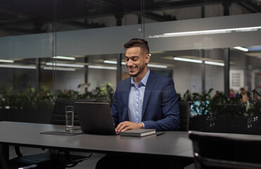 Positive young arabic manager working on laptop at modern hi-tech office, sitting at table, typing...