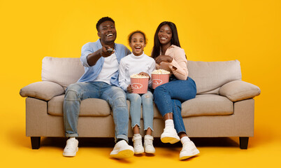 Happy young black family of three people watching movie, parents sitting with daughter on sofa,...