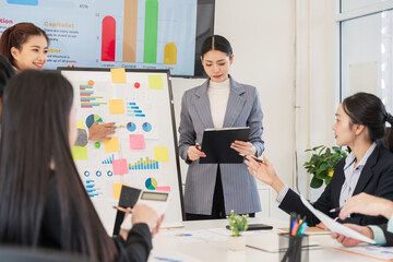 Group of Asian businesswomen presenting their work while their team listens to the explanation. They use laptops to check documents. They have tablets.
