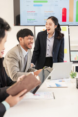 Asian business group discusses new project documents and presents in conference room, laughing and enjoying working together.