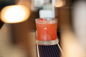 A vibrant red fruity cocktail with pomegranate garnish served in a rocks glass on a bar mat counter, with warm blurred bokeh lights in the foreground background of a modern bar.