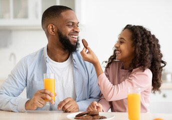 Have A Bite. Portrait of African American little girl feeding her happy daddy with homemade cookie. Smiling black man and his daughter having breakfast at home in the kitchen, drinking orange juice