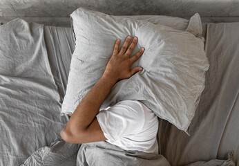 Closeup of irritated young black man lying in bed and covering head and ears with pillow, hearing and suffering from too loud sound. Angry millennial guy can't sleep, tired of noisy neighbors