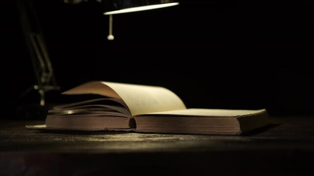 Pages of old book turning under light of table lamp on black background close-up