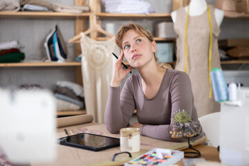 Calm and relaxed young female fashion designer sitting at work table with mug of coffee and tablet in sewing studio, reflecting on new design while browsing references and sketches. Creative process