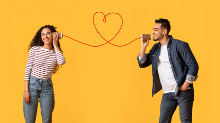 Message With Love. Happy Young Arab Man Telling Romantic Words To His Girlfriend Through Tin Phone With Drawn Red Heart As String, Loving Middle Eastern Couple Posing Over Yellow Background