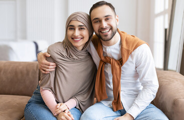 Happy Muslim Couple Embracing Smiling To Camera Sitting Together On Couch At Home. Loving Arab Husband Hugging Beautiful Wife In Hijab. Family, Love And Relationship Concept