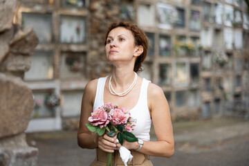 Fototapeta premium Elegant grieving woman in forties, wearing white tank top and pearls, standing with bouquet in hands near stone columbarium wall in cemetery, gazing sorrowfully and contemplating loss